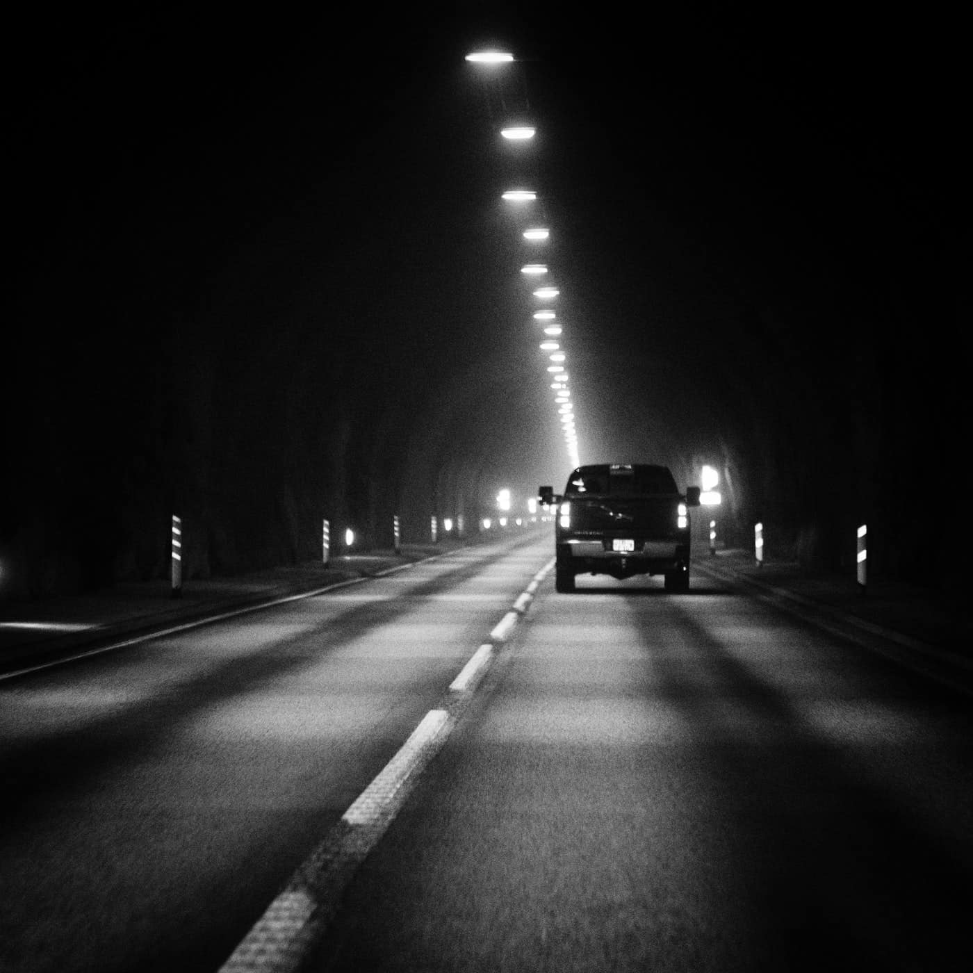 Black and white image of truck driving in a dark tunnel with lights down the middle of the ceiling