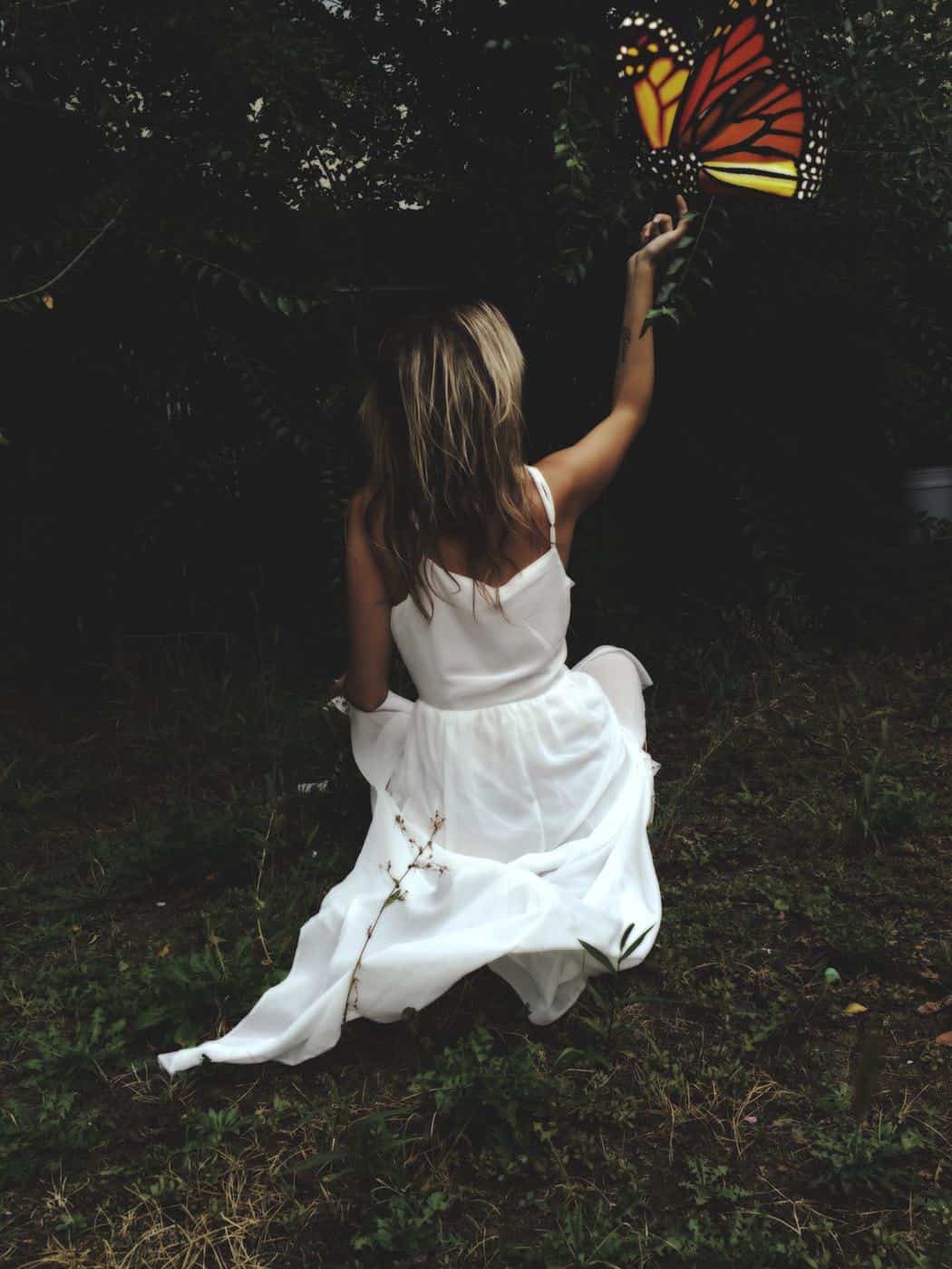 Woman in white dress kneeling in grassy garden stretching her arm to touch a large fantasy butterly