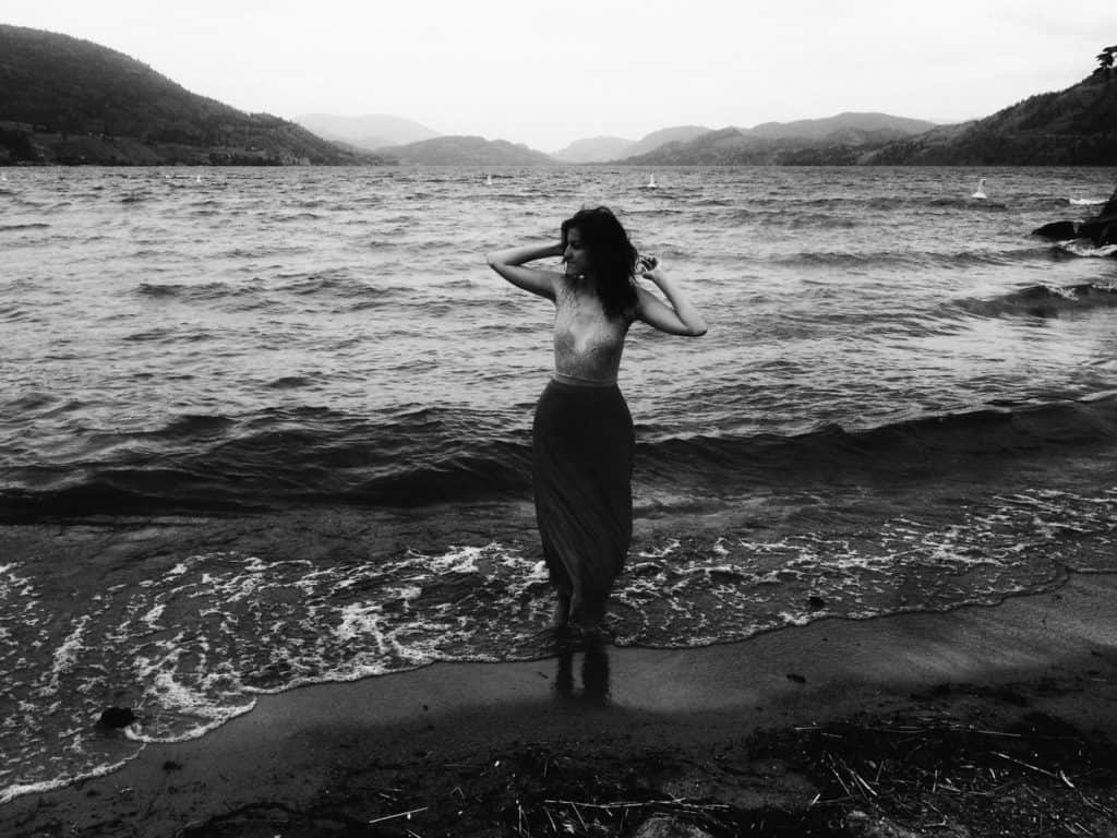 Black and white photo of woman standing beside ocean waves with mountains in the background