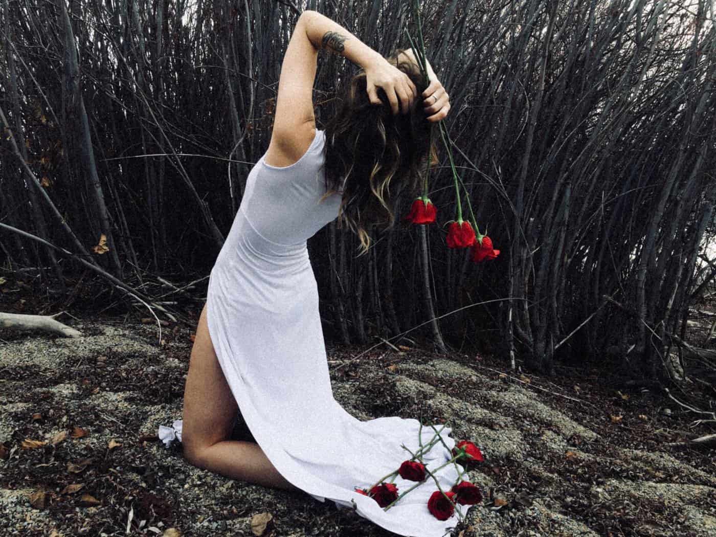 Woman facing away on her knees in white dress with heads on her head and roses in her hands and on ground with bare trees and ground
