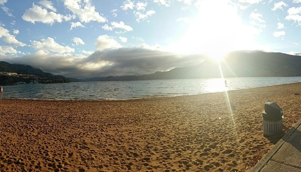 Wide photo of beach and ocean with clouds and mountains in the background along with bright sunflare