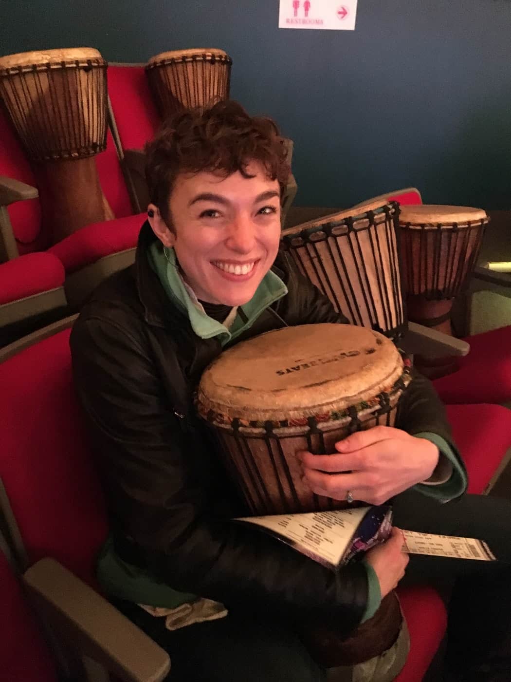 Smiling young woman holding a bongo drum in theatre seats with drums all around