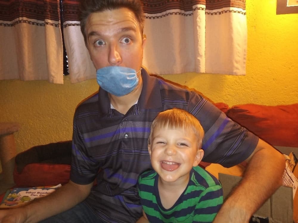 Photo of man wearing medical mask sitting on couch with young son smiling