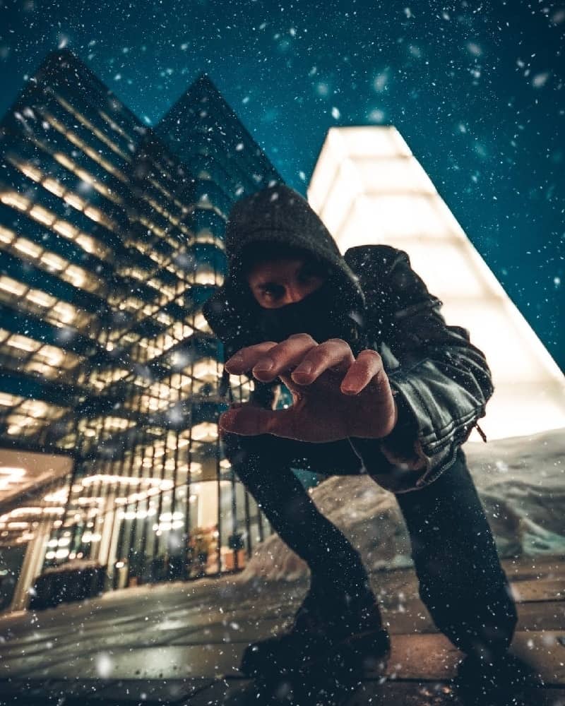Young man with hood and mask reaching down at viewer with city, snow, night sky and building in the background